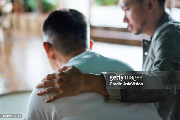 rear view of son and elderly father sitting together at home. son caring for his father, putting hand on his shoulder, comforting and consoling him. family love, bonding, care and confidence - assistance stockfoto's en -beelden