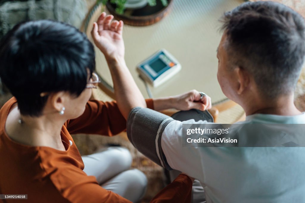 High angle view of senior Asian couple checking blood pressure at home. Wife examining blood pressure on her husband's arm with a blood pressure monitor. Elderly and healthcare concept
