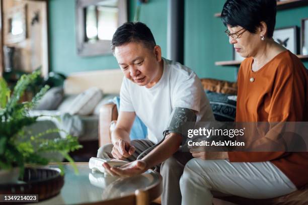 älteres asiatisches paar, das den blutdruck zu hause überprüft. ehefrau untersucht den blutdruck am arm ihres mannes mit einem blutdruckmessgerät. senioren- und gesundheitskonzept - hypertension stock-fotos und bilder