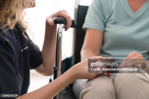 young home nurse holding hands of senior woman in wheelchair. - consoling stock pictures, royalty-free photos & images