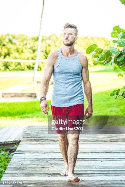 fit young muscular guy walks on a beach boardwalk outdoors in coastal location - swimwear singlet stock pictures, royalty-free photos & images