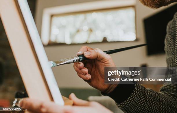 a man wearing glasses concentrates as he paints with oils onto a canvas on an easel - pittore artista foto e immagini stock