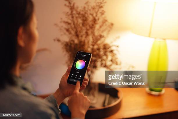 young woman using smartphone to adjust the lighting equipment of a modern smart home - smart homes stock pictures, royalty-free photos & images