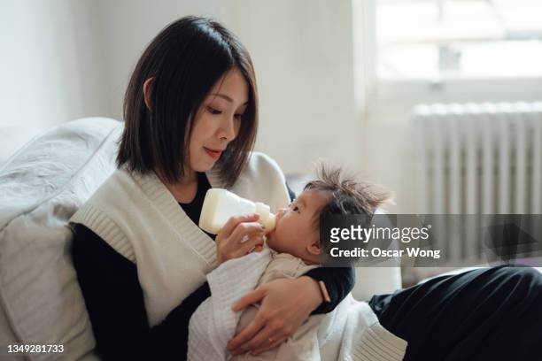 mother feeding baby with bottle on sofa - baby bottle stockfoto's en -beelden