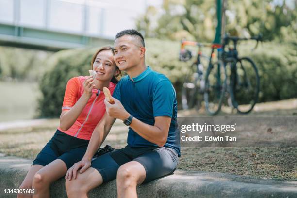 asian chinese couple cyclist taking a break enjoying sandwiches at river front after morning road trip cycling in public park - cycling vest stock pictures, royalty-free photos & images