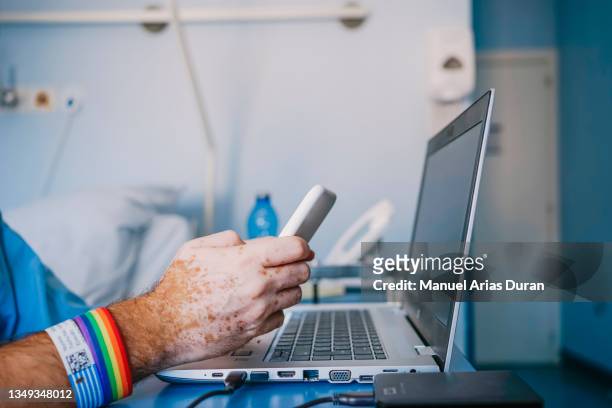 close-up of the hands of a patient in the hospital using his laptop and cell phone. the patient is wearing a lgtb bracelet. - cellphone cancer illness stock-fotos und bilder