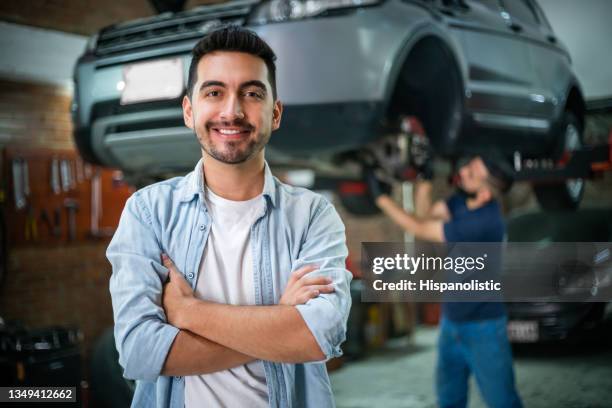 satisfied male customer at the car garage facing camera with arms crossed while mechanic checks the wheels of the car at background - auto repair shop background stock pictures, royalty-free photos & images