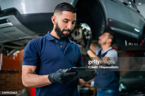 supervisor in einer autowerkstatt, die das tablet überprüft, während der mechaniker im hintergrund an einem auto arbeitet - auto tablet stock-fotos und bilder