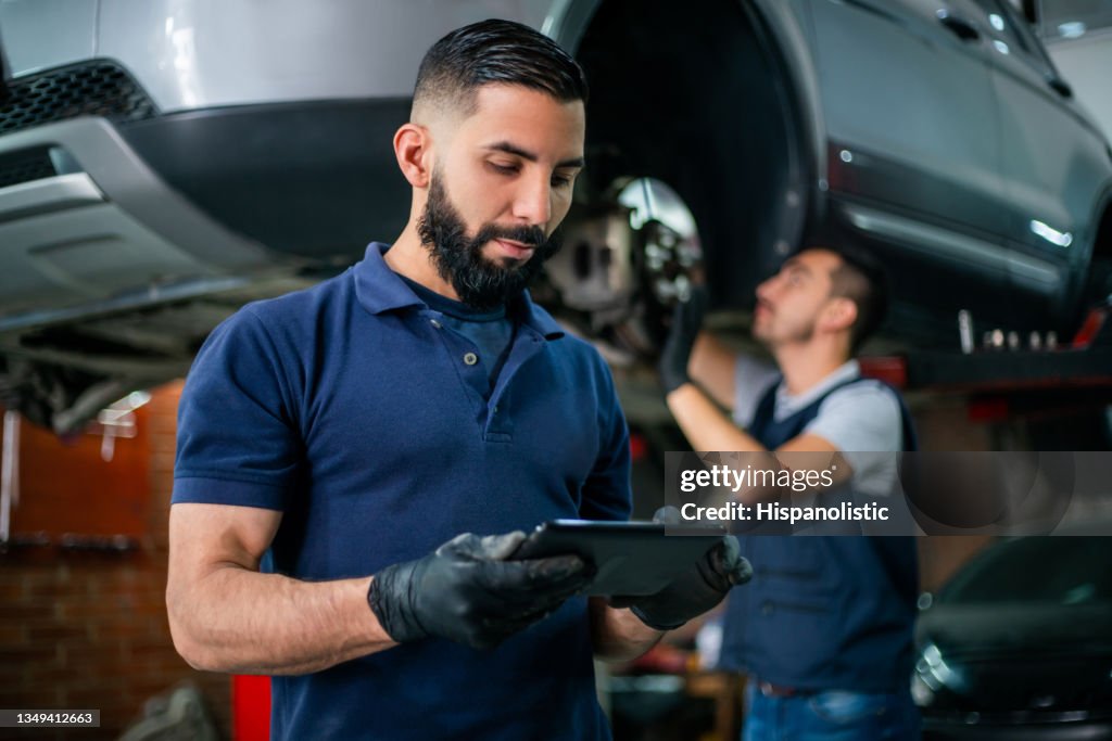 Supervisor at a car workshop checking tablet while mechanic works at background on a car