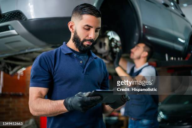 supervisor at a car workshop checking tablet while mechanic works at background on a car - machine part stock pictures, royalty-free photos & images