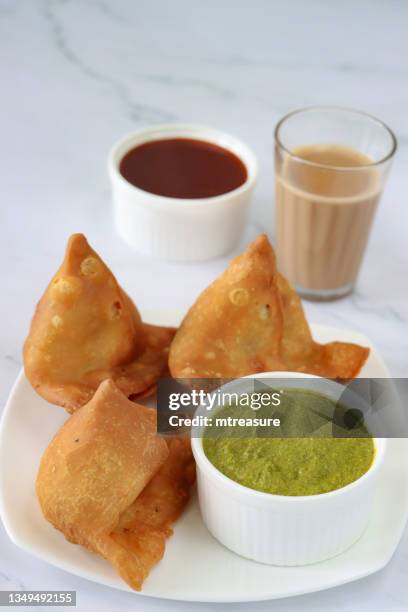 nahaufnahme von drei gebratenen samosas auf einem weißen teller, gefüllt mit gewürzten kartoffeln, erbsen und fleisch neben ramekins aus minzkoriander-dip und mango-chutney, glas chai-tee, marmoreffekthintergrund, erhöhte ansicht, fokus auf vordergrund - chutney stock-fotos und bilder