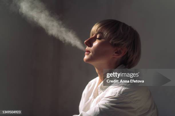 modern tattooed woman with a short blond hair is smoking a e-cigarette indoors. she is wearing a light casual unisex closing. concept of gender neutrality - e zigarette stock-fotos und bilder