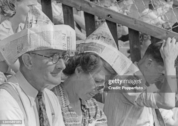 Two spectators wear paper hats made from newspapers sitting alongside a man scratching his head as they attempt to keep cool in the high temperatures...