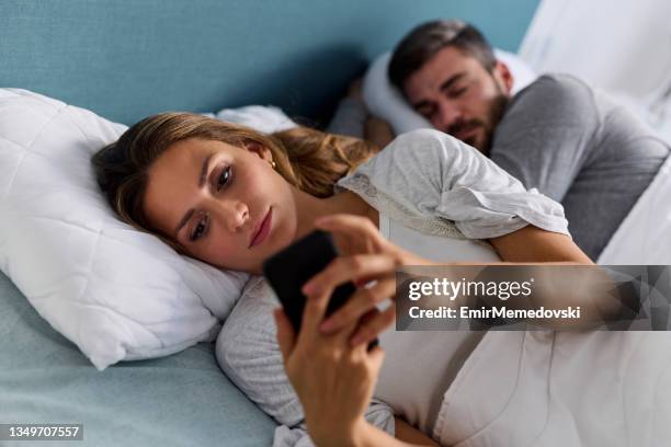 young woman using mobile phone and checking messages in bed while her husband is asleep - affari stock pictures, royalty-free photos & images