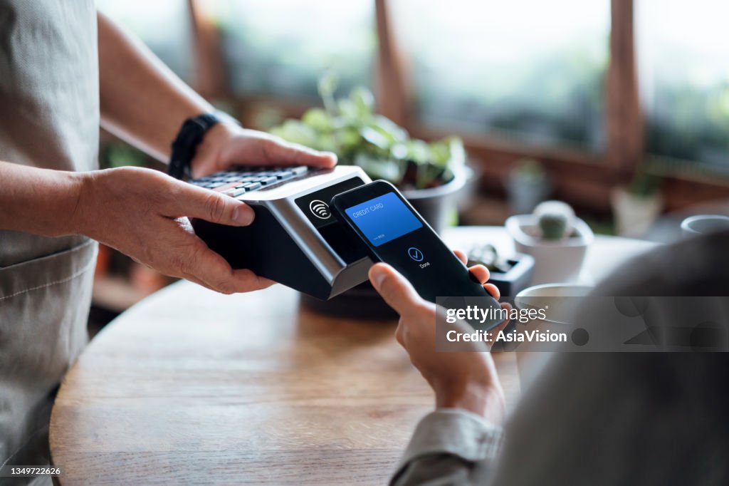 Nahaufnahme der Hand eines Mannes, der die Rechnung mit Kreditkarte bezahlt, kontaktlos auf dem Smartphone in einem Café, scannend auf einem Kartenautomaten. Elektronische Zahlung. Banking und Technologie