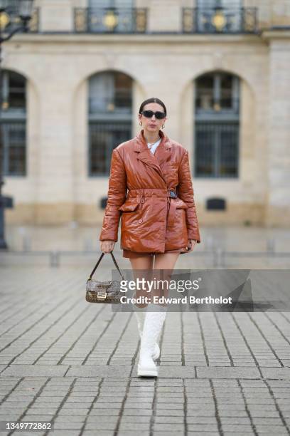 Gabriella Berdugo wears black sunglasses, gold earrings, a white t-shirt, a gold and silver necklace, a brown shiny leather belted puffer jacket,...