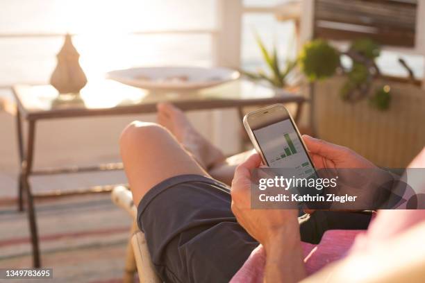 man checking financial trading data on mobile phone while relaxing on terrace, close-up of hands - etf fotografías e imágenes de stock