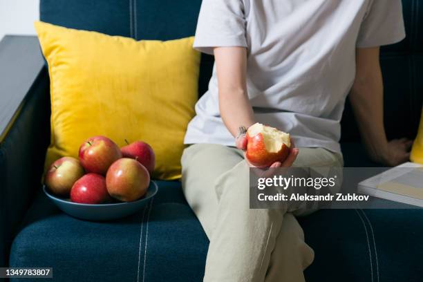 a red apple in the hands of a woman who is sitting on a bright-colored sofa at home. the girl eats a fruit, bites off a piece of juicy apple. cultivation of organic farm products. the concept of vegetarian, vegan, raw food and diet. - pomme croquée photos et images de collection