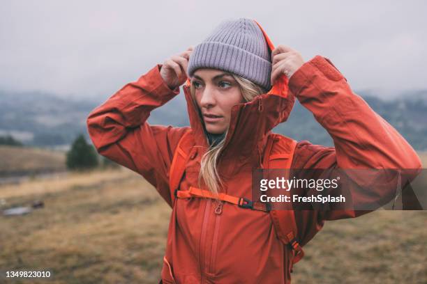 porträt einer schönen blonden wandererin in roter jacke und grauem hut, die an einem bewölkten tag die berge erkundet - jacket stock-fotos und bilder
