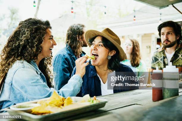 medium shot of laughing woman feeding friend chip with guacamole while eating at food truck - toma mediana fotografías e imágenes de stock