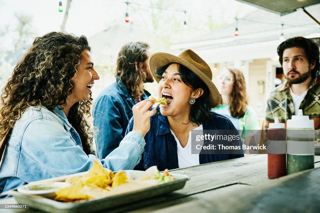 Medium shot of laughing woman feeding friend chip with guacamole while eating at food truck