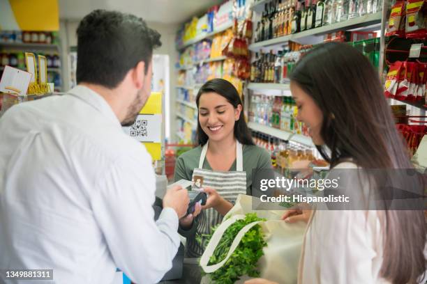 latin american couple paying for their groceries at a small market with credit card while friendly cashier holds the credit card reader - latin american and hispanic shopping bags bildbanksfoton och bilder