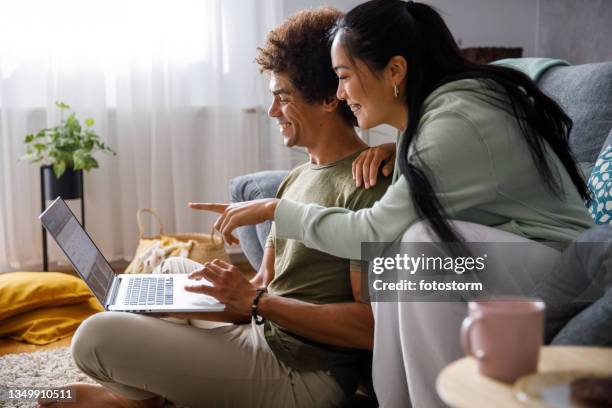 young woman pointing at something on laptop screen while looking at online banking with her boyfriend - buying online stock pictures, royalty-free photos & images