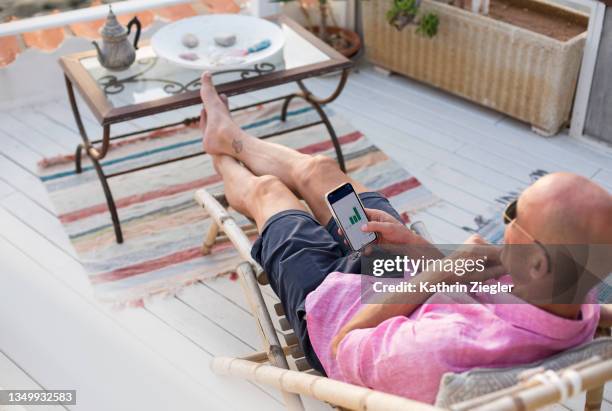 man checking financial trading data on mobile phone while relaxing on deck chair - early retirement stockfoto's en -beelden
