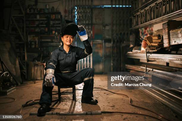 saldatrice femminile asiatica cinese sorridente con abbigliamento da lavoro protettivo garage seduto sullo sgabello nel laboratorio - fabbro ferraio foto e immagini stock