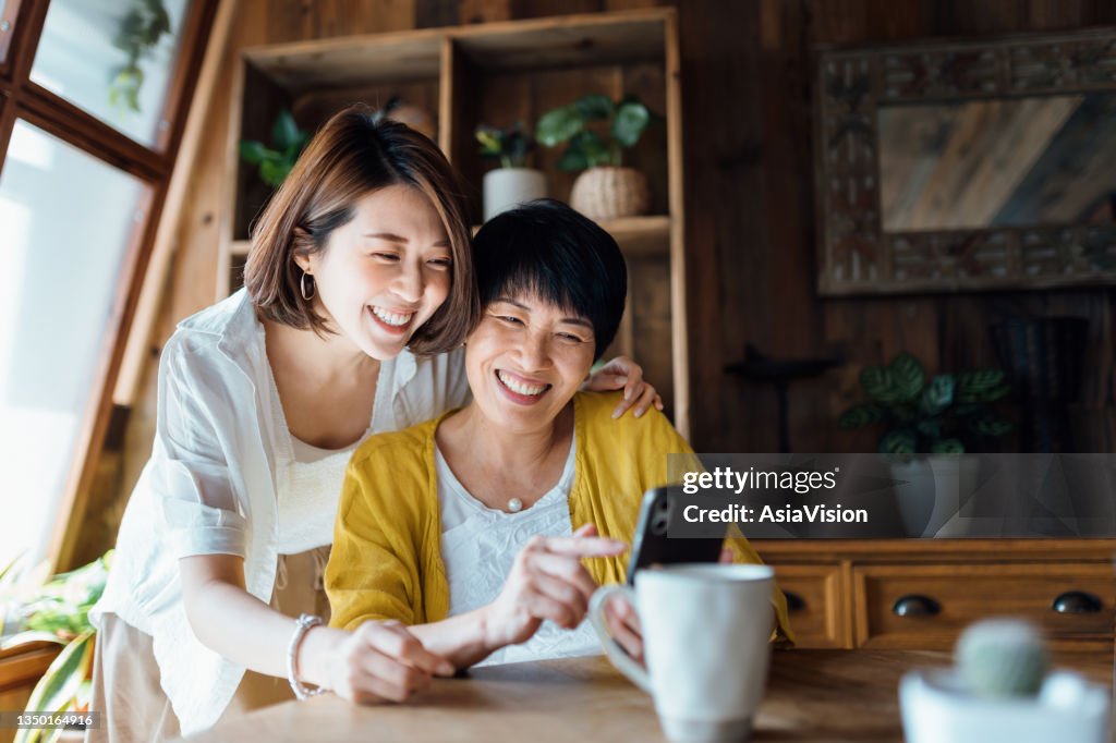 Liebevolle asiatische Seniorin Mutter und Tochter nutzen das Smartphone gemeinsam zu Hause, lächeln fröhlich, genießen Mutter und Tochter Bindungszeit. Mehrgenerationenfamilie und Technologie