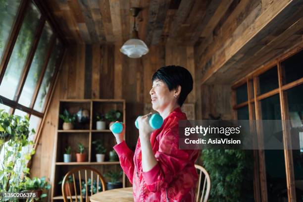 active senior asian woman exercising at home. practicing fitness training and lifting dumbbells. maintaining healthy fitness habits. elderly wellbeing, health and wellness concepts - elderly cognitive stimulation therapy stockfoto's en -beelden