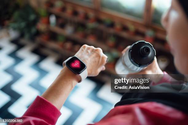 over the shoulder view of senior asian woman resting after exercising at home, looking at her smart watch, using fitness tracker app and measuring pulse. maintaining healthy fitness habits. elderly wellbeing, health, wellness and technology concept - wearable computer bildbanksfoton och bilder