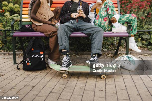 low section of multiracial friends sitting on bench at park - parkbank stock-fotos und bilder