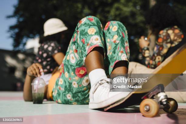 young non-binary person lying down with skateboard and friends in park during sunny day - laufschuhe stock-fotos und bilder