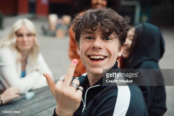 portrait of happy teenage boy doing obscene gesture while sitting with friends - v sign stock pictures, royalty-free photos & images