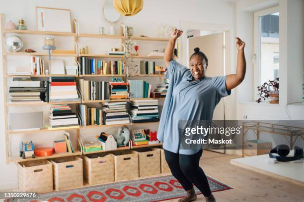 joyful mid adult woman dancing with arms raised at home - frau tanzt im wohnzimmer stock-fotos und bilder