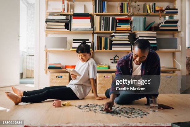 man playing jigsaw puzzle while girlfriend reading book in living room - puzzle stock-fotos und bilder