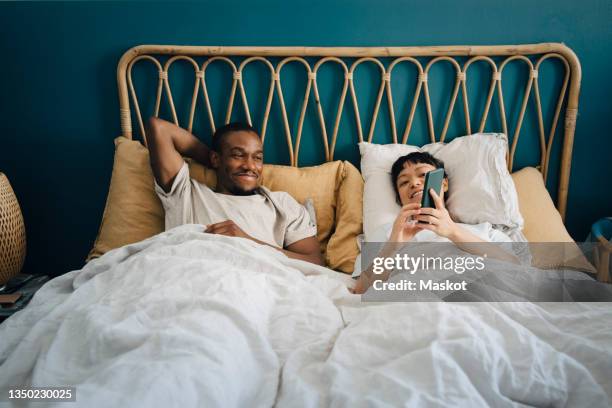 smiling girlfriend sharing smart phone with boyfriend while lying on bed - couple in bed stock pictures, royalty-free photos & images