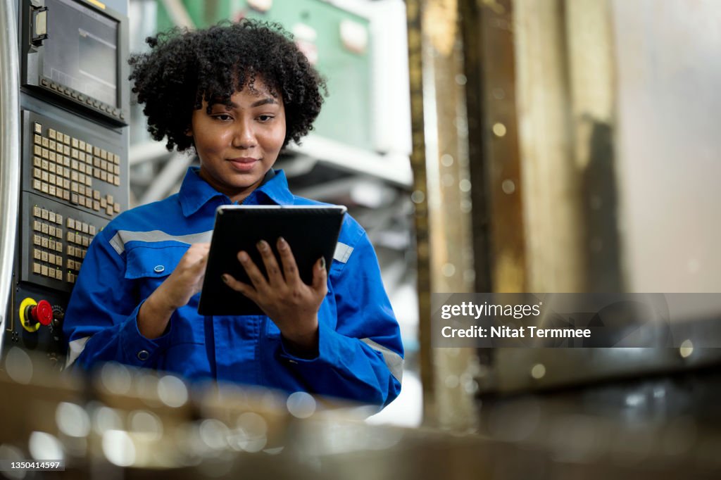 CNC milling machine setup process. Female African American Mechanical Engineer prepares the machine and engineering workpiece base on the machining standard. She is using a tablet in a production line.