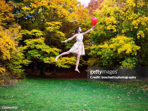 glückliche junge brünette frau, die mit einem roten ballon durch die herbstlandschaft fliegt - alice in wonderland stock-fotos und bilder