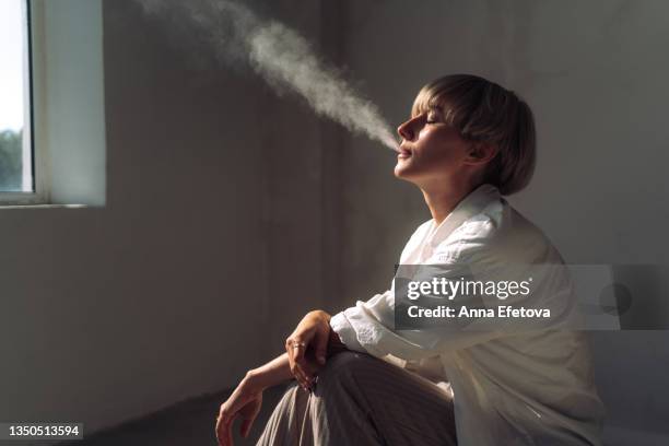 modern woman with a short blond hair is smoking indoors. she is wearing a light casual unisex closing. concept of gender neutrality - vape stock pictures, royalty-free photos & images