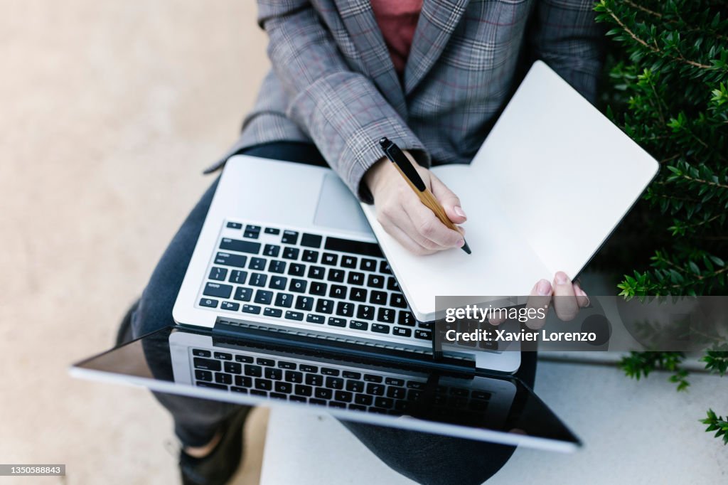 Business woman taking notes in a notebook while working with her laptop outdoors.