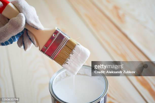 the hands of a woman in protective gloves hold a brush on a wooden background. a trickle of paint drips into a jar of white paint. painting and repair work. the concept of construction, hobby, restoration. - emaille stock-fotos und bilder