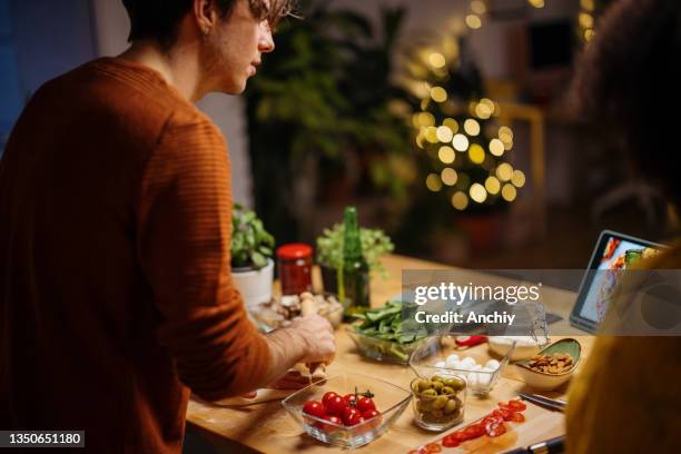 young man looking at digital tablet and talking to his girlfriend while preparing pizza for dinner - vegetarian christmas dinner stock pictures, royalty-free photos & images