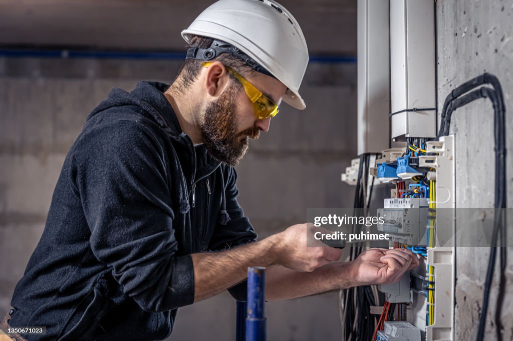 A male electrician works in a switchboard with an electrical connecting cable.