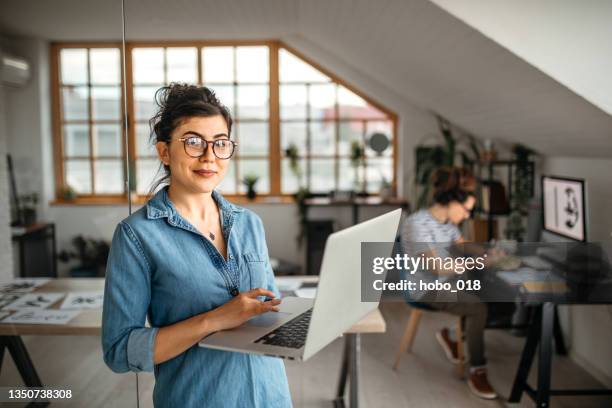 カメラを見てラップトップを持つ若いサラリーマンの女性 - creative desk ストックフォトと画像