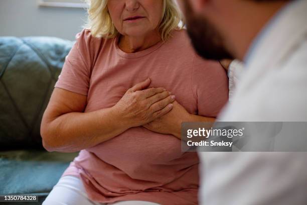 senior woman is sitting and holding her chest during doctor's appointment.  a young intern is talking to his patient. selective focus. - human heart bildbanksfoton och bilder
