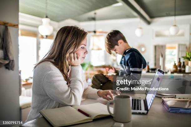 woman working from home on laptop while son uses smartphone - kitchen coffee home stock-fotos und bilder