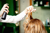 Female coiffeur fixing hairstyling of blonde woman with hairspray in a beauty salon.