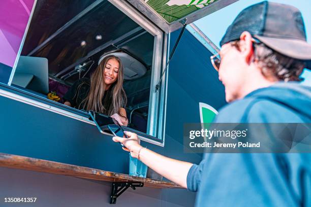 young female business owner standing giving credit card reader to a customer to use a smartphone to pay for goods using nfc cashless technology at a food truck concession stand - food truck payments stock pictures, royalty-free photos & images